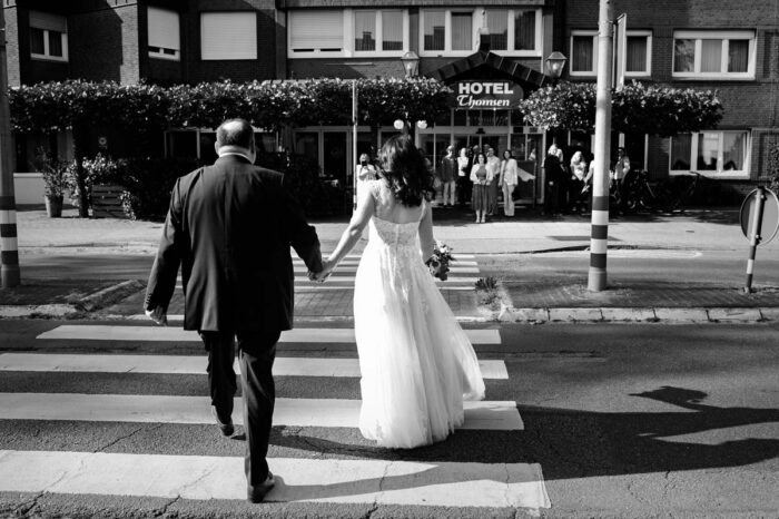 braut und Bräutigam gehen Hand in Hand über die Straße zur hochzeitslocation, während ihre gaste im Hintergrund auf sie warten.